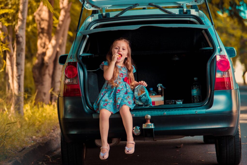 little girl sat in the back of a family car, eating a snack.