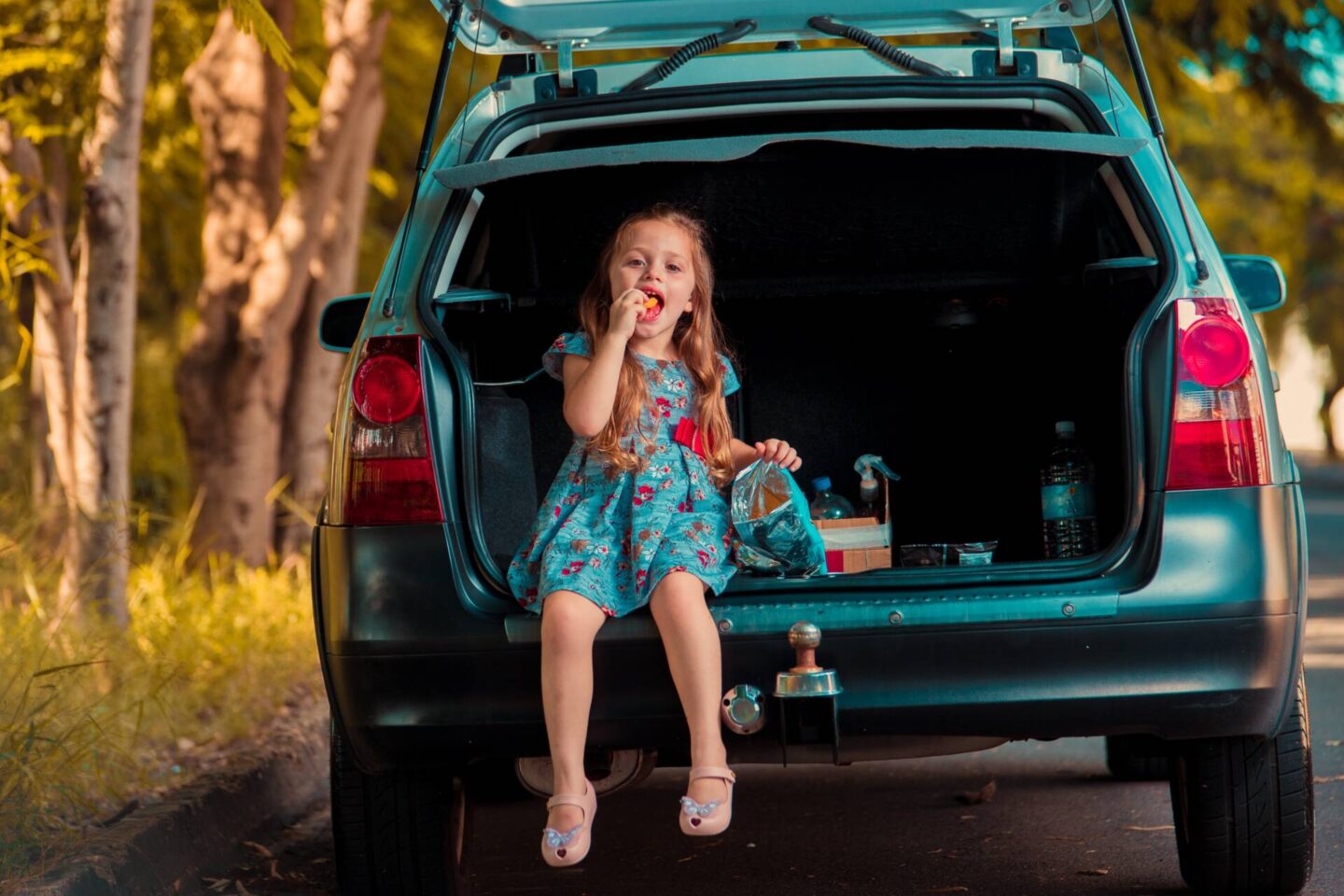 little girl sat in the back of a family car, eating a snack.