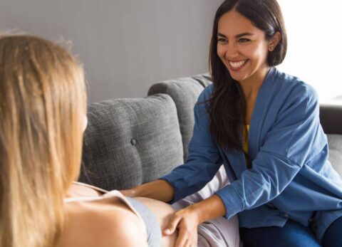 community midwife in plain clothes feeling the pregnant abdomen of a lady reclining on a couch in her home