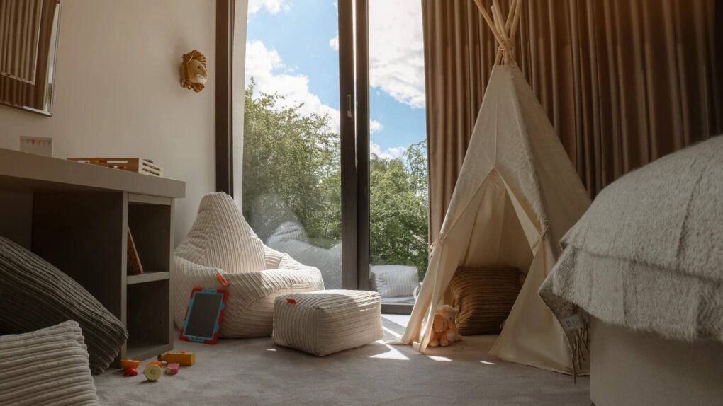 a childs bedroom with large windows, a bean bag and footstool, wigwam and a lion's head on the wall. Some wooden blocks are on the floor.
