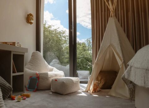 a childs bedroom with large windows, a bean bag and footstool, wigwam and a lion's head on the wall. Some wooden blocks are on the floor.