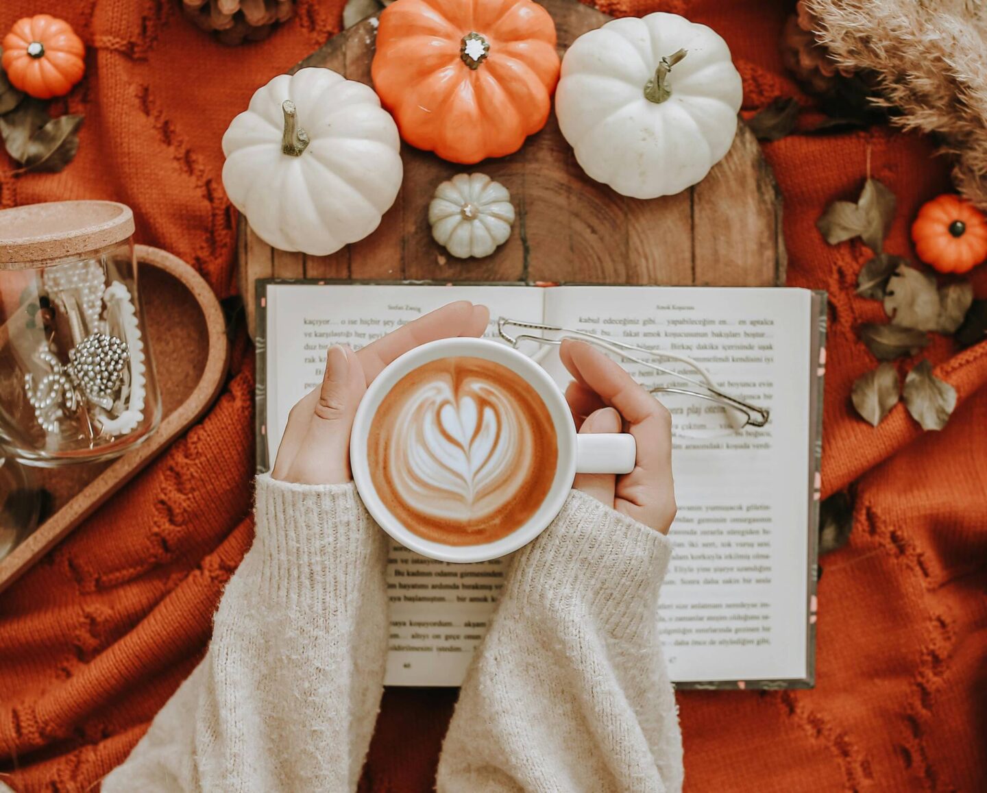 womans hands and arms wearing a cream sweater holding a cup of coffee on top of a book and blanket, there are white and orange pumpkins