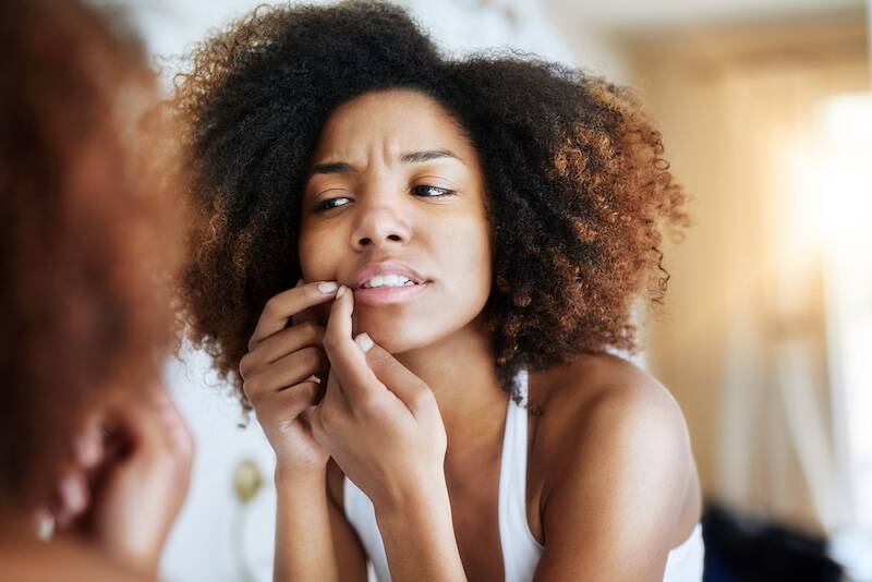 Woman, mirror and acne with skincare in home with pimple breakout, skin problem and frustrated. African girl, reflection or face inspection in bathroom for beauty hygiene, pop blackhead and self care.